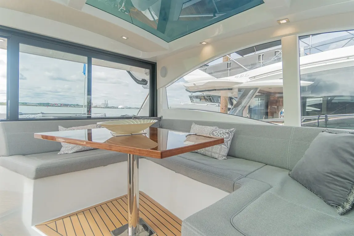 Luxury yacht interior with grey seating, a wooden table, and panoramic windows overlooking the marina and waterfront scenery.